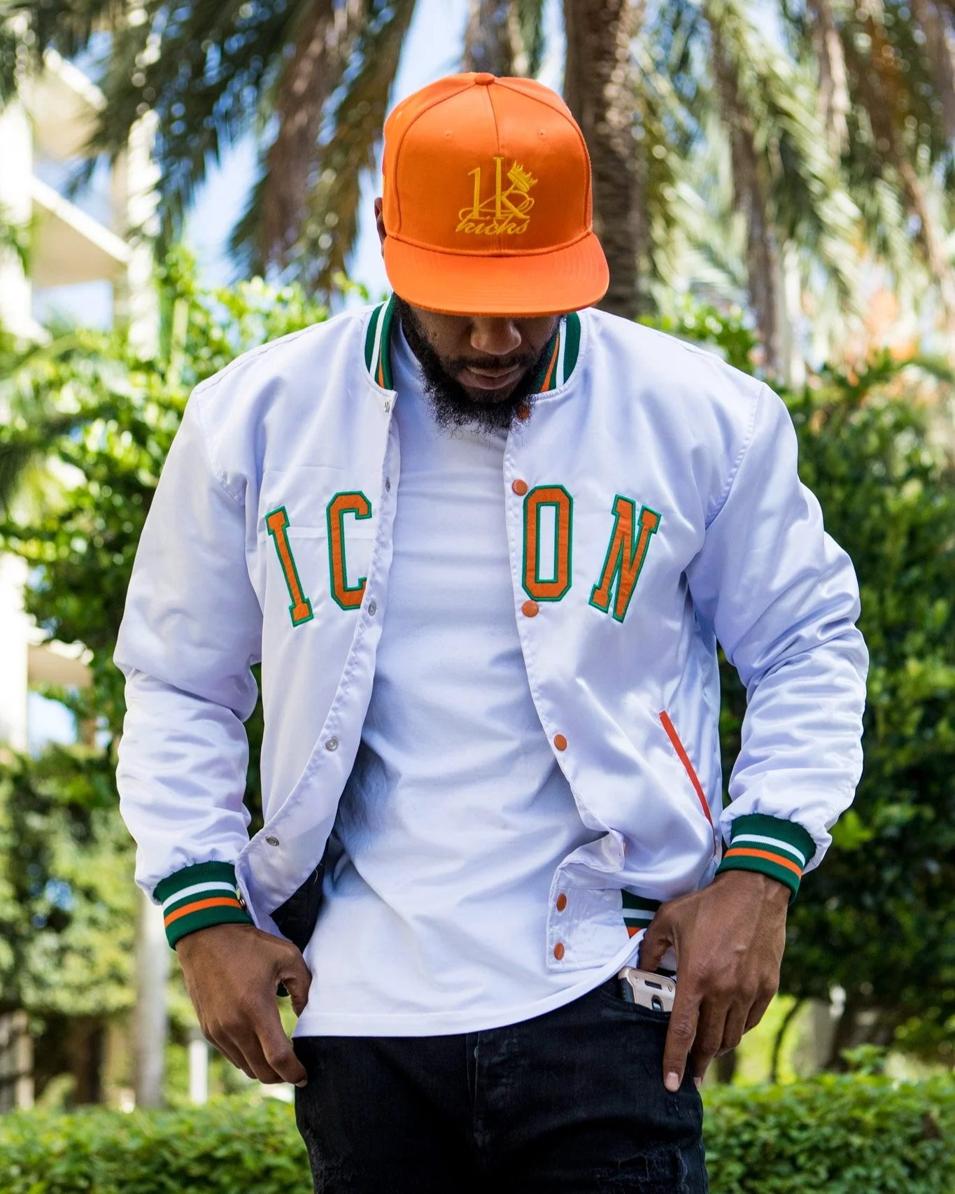 Man wearing 1K Kicks white letterman jacket with green and orange accents, an orange cap, and black pants outdoors.