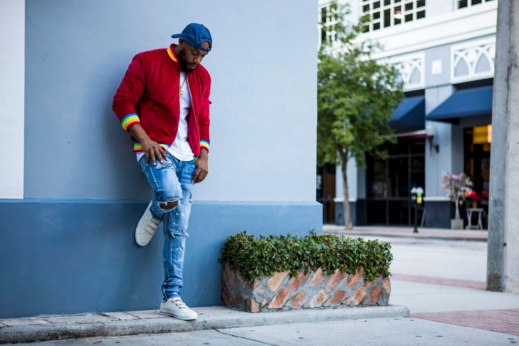 Model leaning against a wall wearing the 1K Spectrum red suede streetwear jacket in a sunny Florida city setting